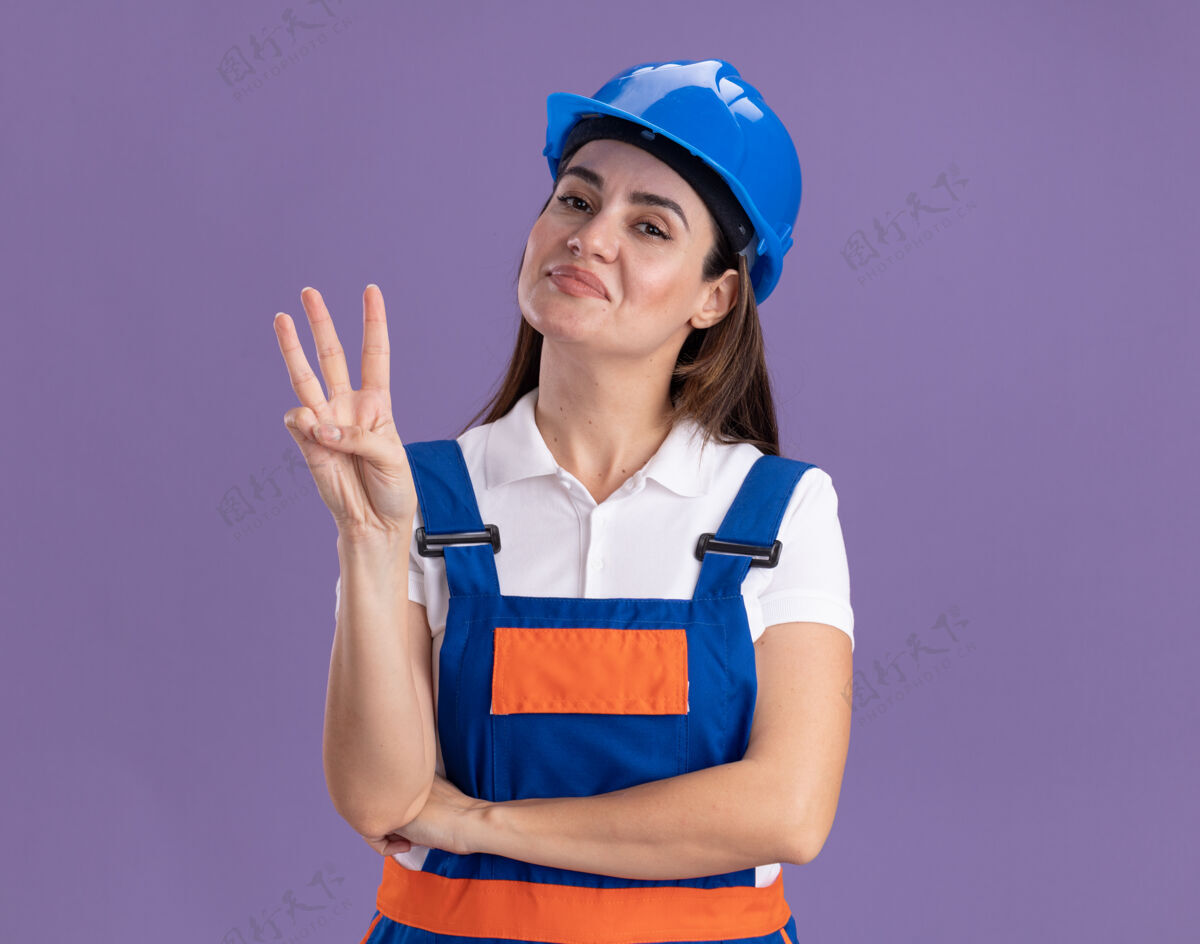 建筑工人身着制服的年轻建筑妇女高兴地在紫色的墙上显示三个孤立的拜托表演年轻