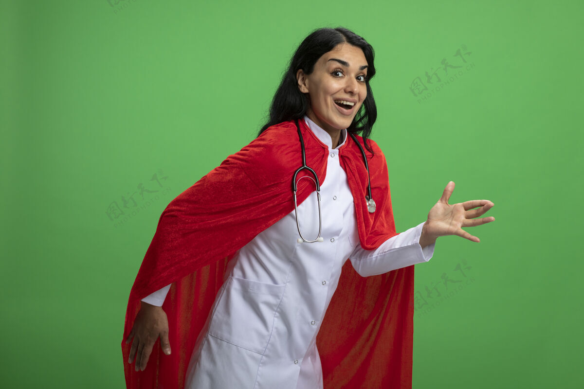 超级英雄快乐的年轻超级英雄女孩穿着医用长袍和听诊器显示机器人风格隔离在绿色医疗机器人表演