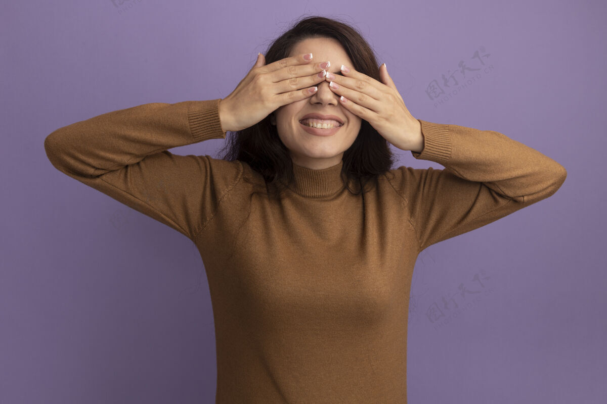 毛衣微笑的年轻漂亮女孩穿着棕色高领毛衣 眼睛蒙着 手孤立在紫色的墙上穿微笑手