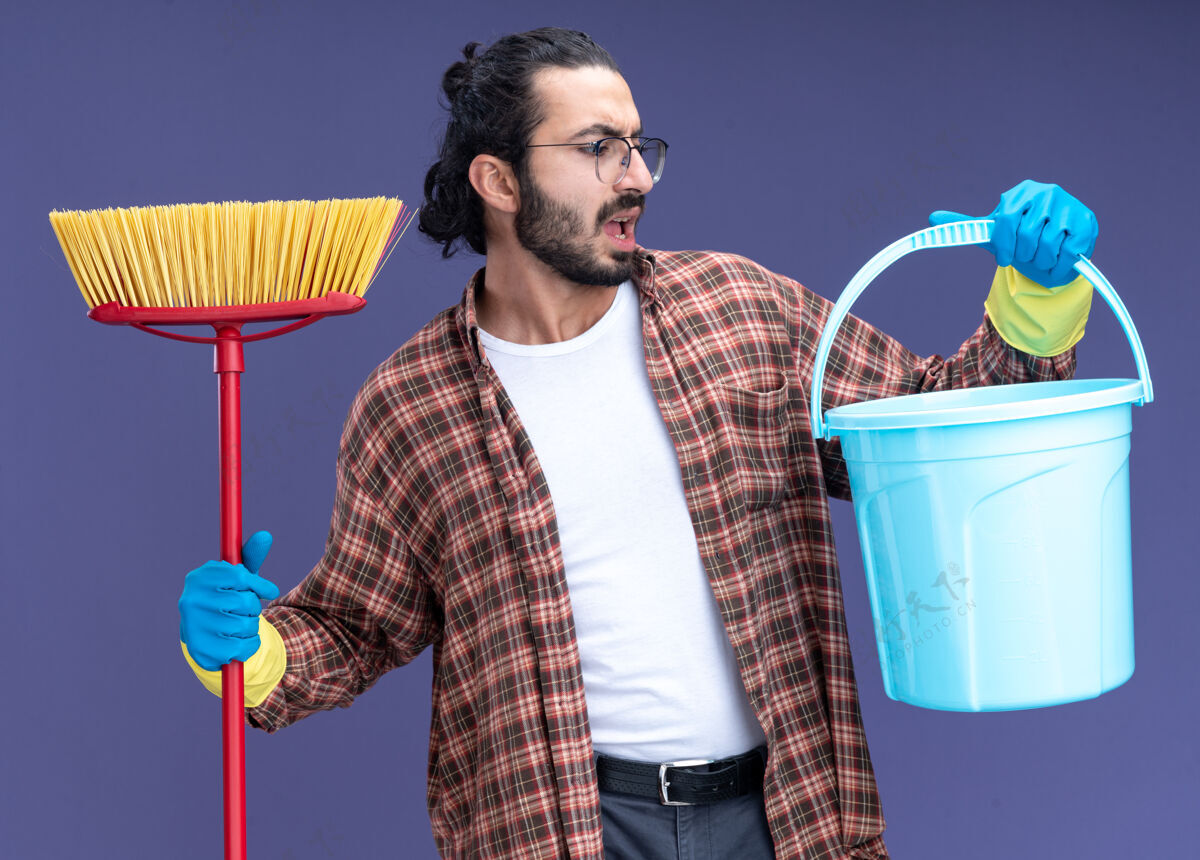 家伙年轻帅气的清洁工穿着t恤 戴着手套 拿着拖把 看着他手上的水桶 孤零零地站在蓝色的墙上拿着桶拖把