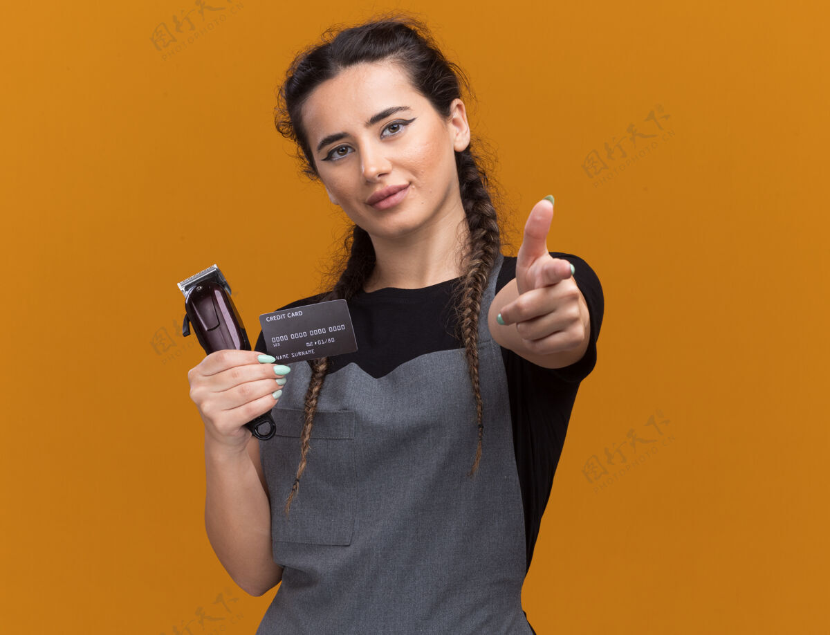 剪子高兴的年轻女理发师穿着制服拿着信用卡和发夹点隔离在橙色的墙上女的制服年轻的