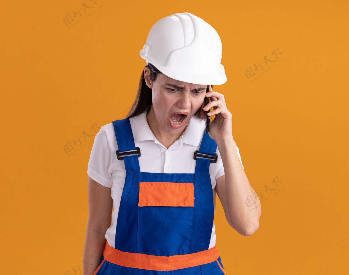 女人愤怒的穿着制服的年轻建筑工人在电话里讲话 被隔离在橙色的墙上年轻制服愤怒