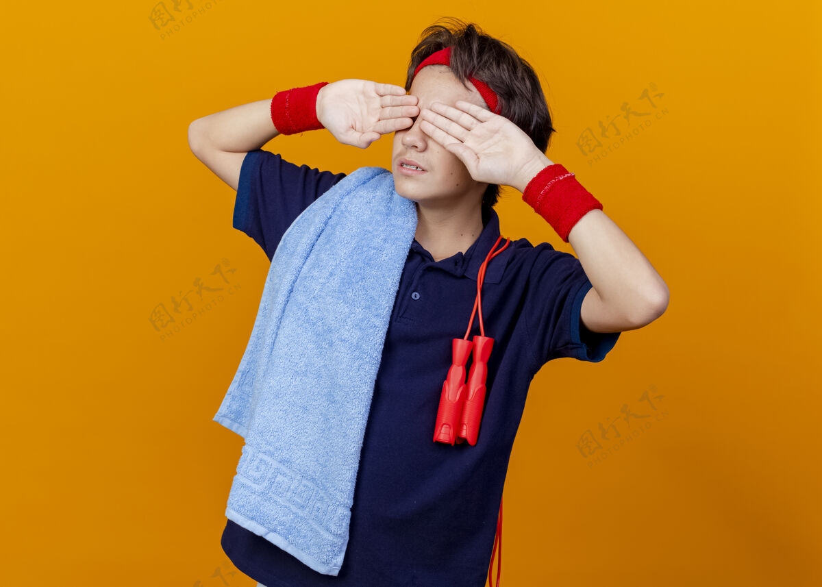 手年轻英俊的运动男孩戴着头带和护腕 戴着牙套和毛巾 肩膀上有跳绳 手放在橙色墙上的眼睛上穿腕带姿势
