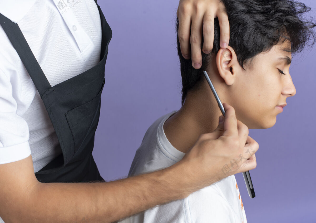 理发年轻帅气的理发师穿着制服在紫色背景上为年轻客户理发年轻穿理发师