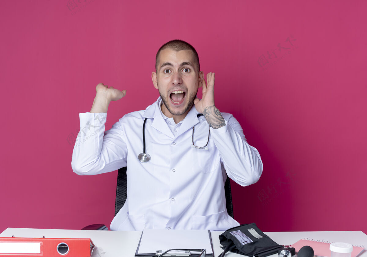 办公桌令人印象深刻的年轻男医生穿着医用长袍和听诊器坐在办公桌前 工作工具显示空手 并保持另一个靠近头部的粉红色隔离年轻工作另一个