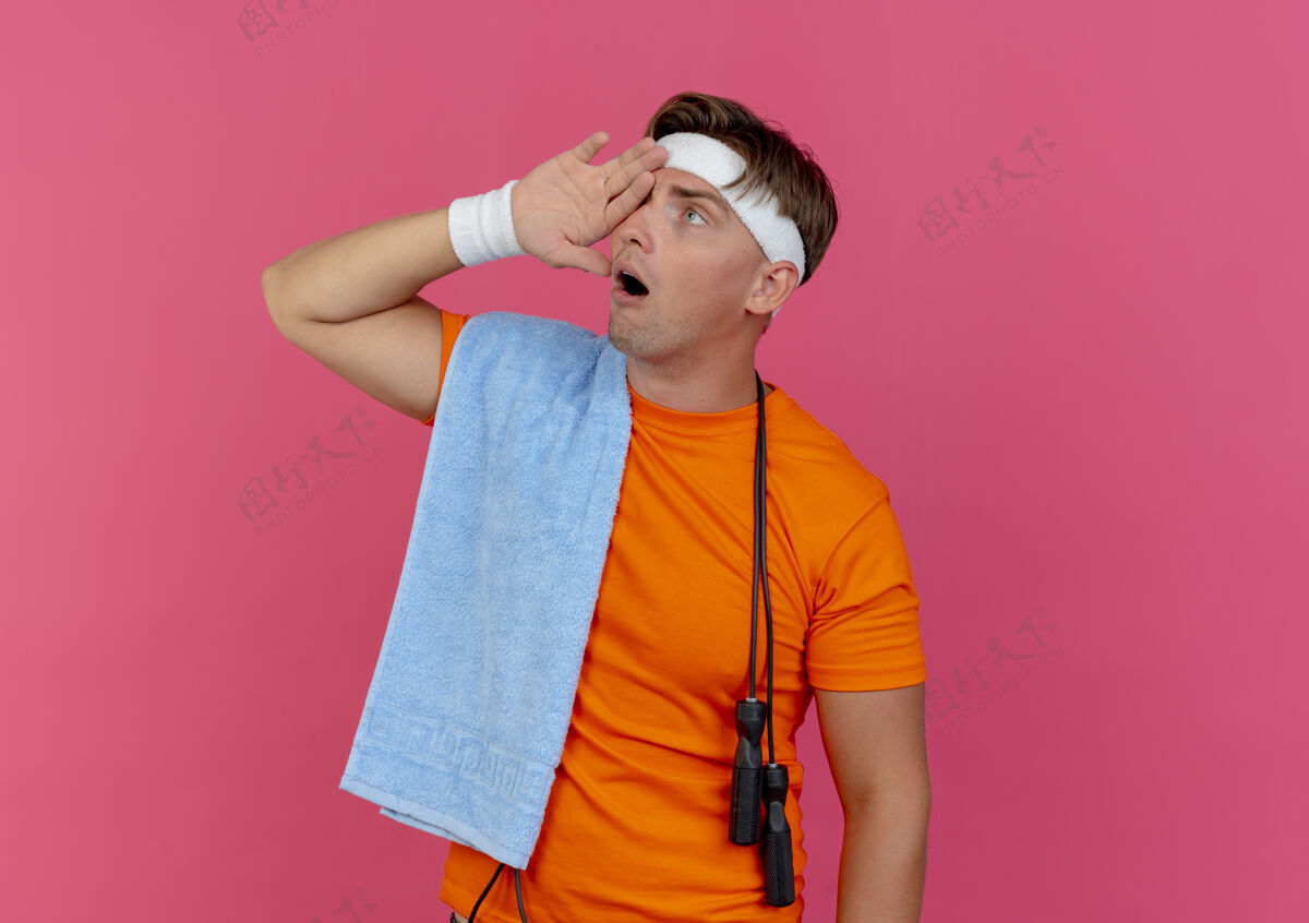 穿印象深刻的年轻英俊的运动男子戴着头带和手镯用毛巾和跳绳围住脖子把手放在眼睛上看着旁边孤立的粉红色脖子印象毛巾