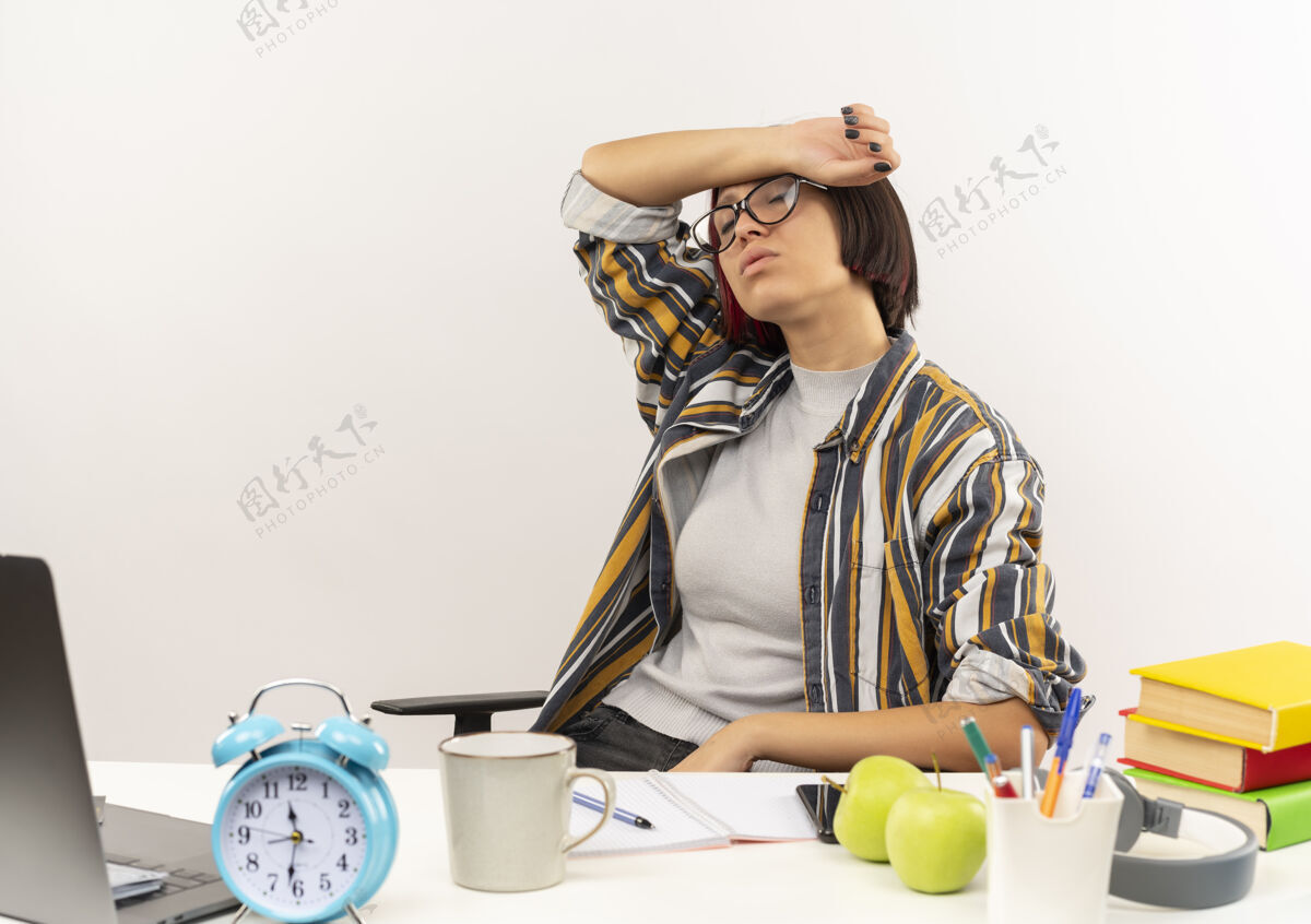 工具疲惫的年轻女生戴着眼镜坐在书桌前用大学工具把胳膊放在额头上 闭着眼睛在白色背景上隔开年轻女孩眼睛