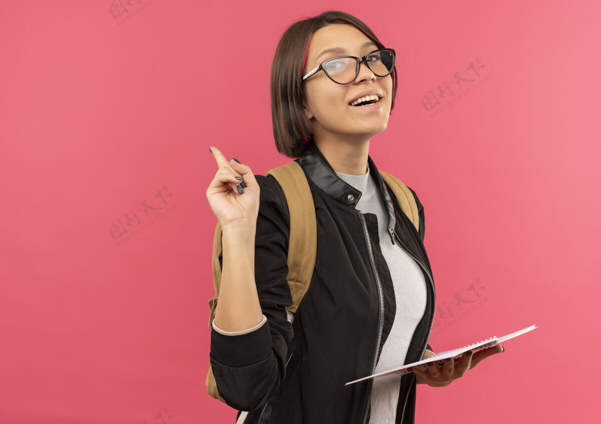 年轻快乐的年轻学生女孩戴着眼镜 背着书包拿着便签本举起手指孤立在粉红色的脸上手指垫包