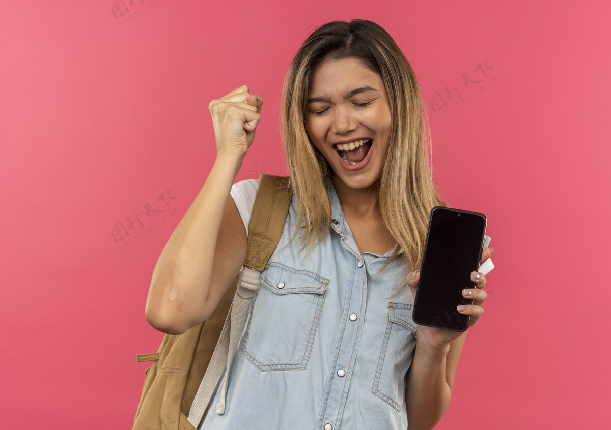 喜悦快乐的年轻漂亮的学生女孩背着书包拿着手机举起拳头闭上眼睛孤立在粉红色学生手机持有
