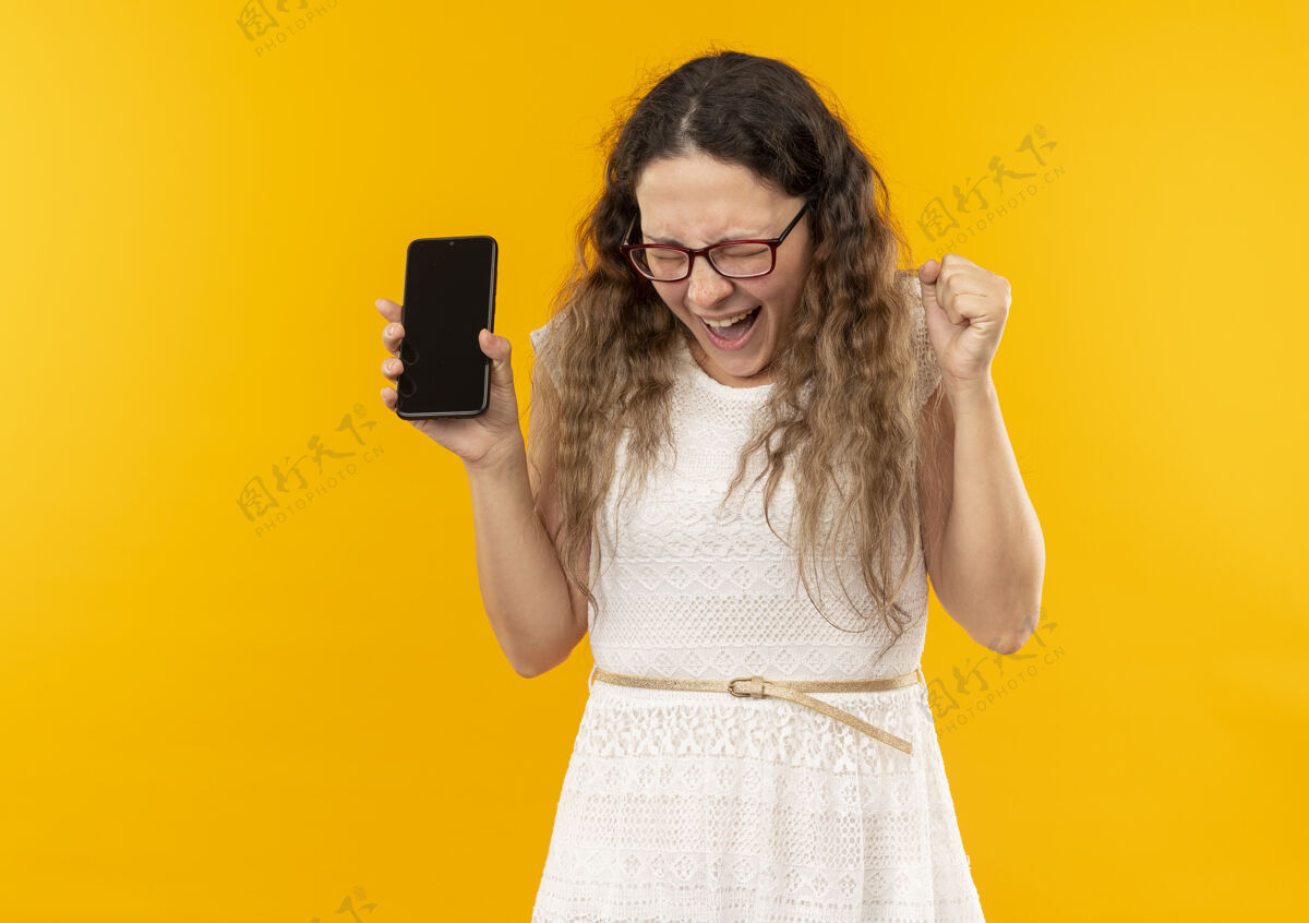 眼镜快乐的年轻漂亮的女学生戴着眼镜背着包拿着手机紧握拳头闭着眼睛孤立在黄色高兴包戴着