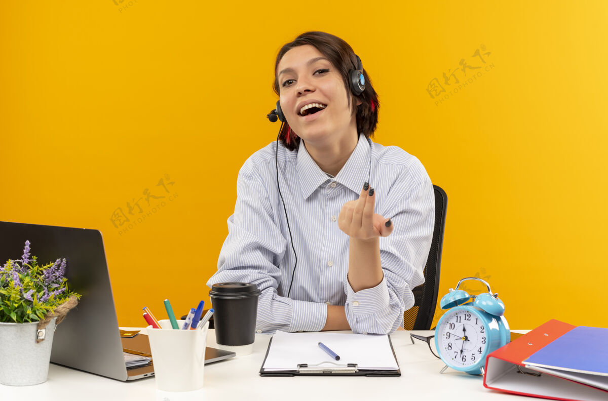 高兴快乐的年轻呼叫中心女孩戴着耳机坐在办公桌旁做着来这里的手势孤立在橙色上穿上耳机电话