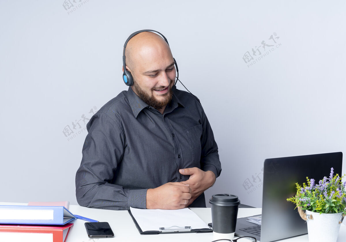 工具高兴的年轻秃头呼叫中心男子戴着耳机坐在办公桌旁 拿着工作工具看着笔记本电脑 手放在肚子上 隔离在白色背景上笔记本电脑穿电话