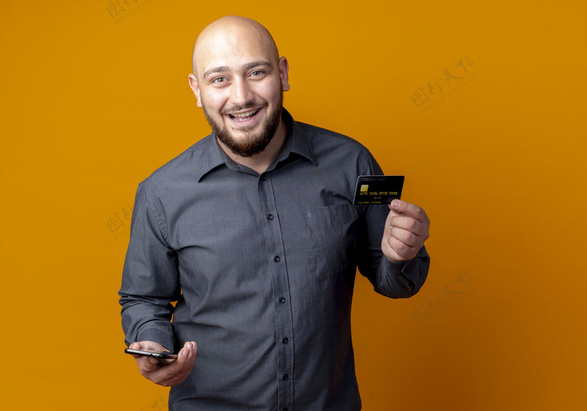秃头快乐的年轻秃头呼叫中心男子手持信用卡和手机孤立在橙色电话年轻手机