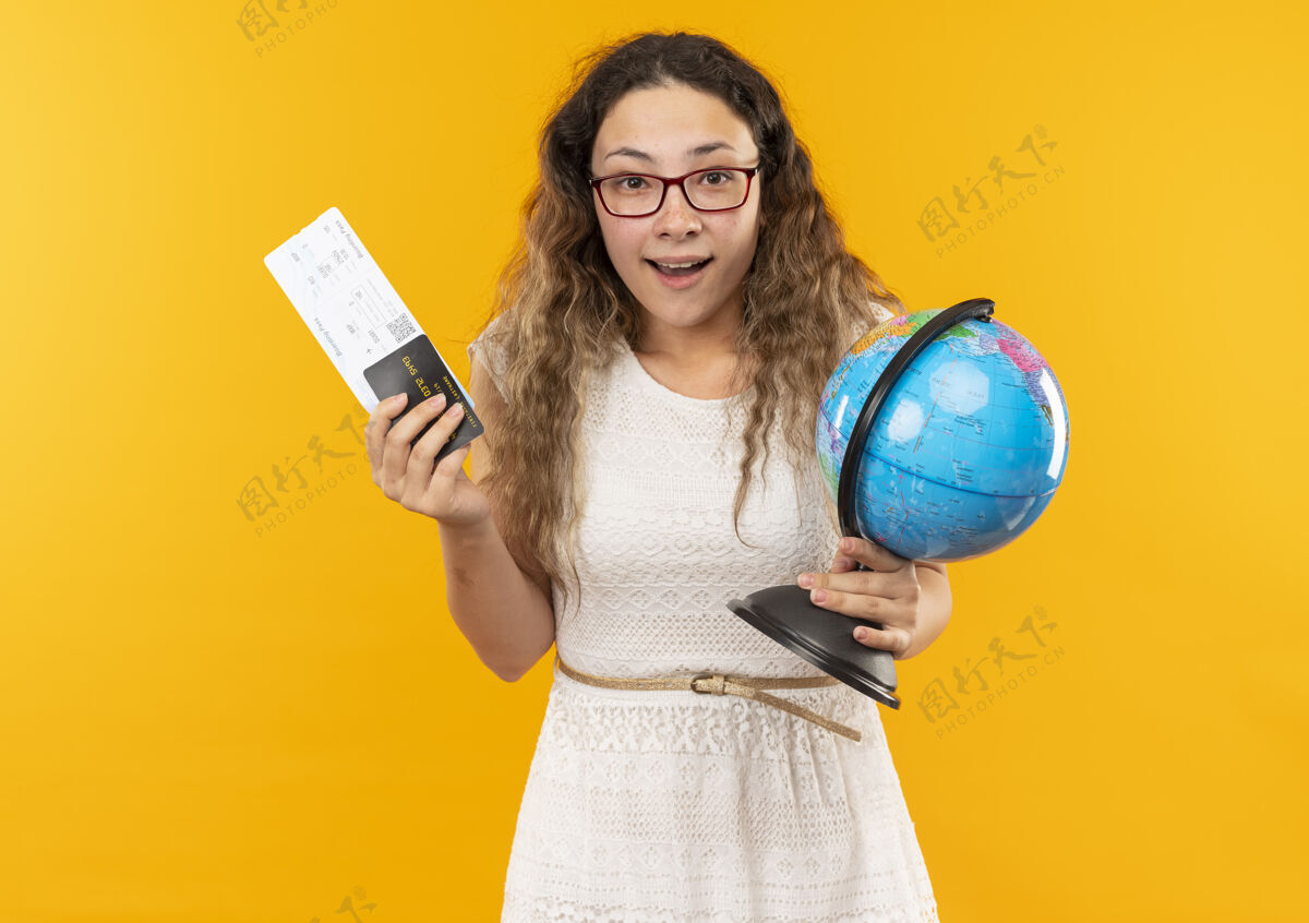 穿着印象深刻的年轻漂亮的女学生戴着眼镜拿着机票 信用卡和地球仪孤立在黄色印象眼镜持有