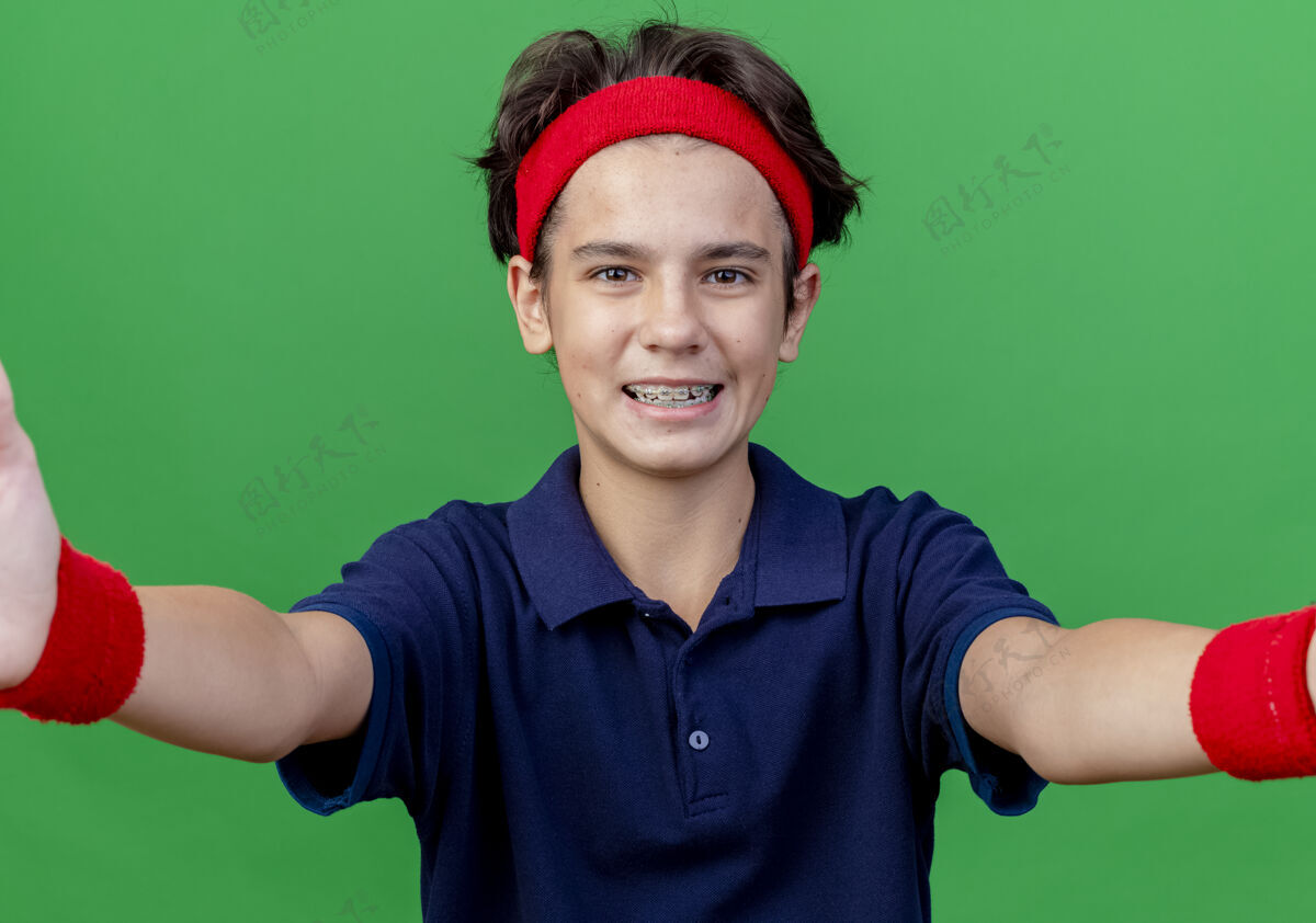 运动快乐的年轻帅气的运动男孩戴着头带和护腕 戴着牙套 看着前面 向前面伸出双手 隔离在绿色的墙上佩戴背带市民