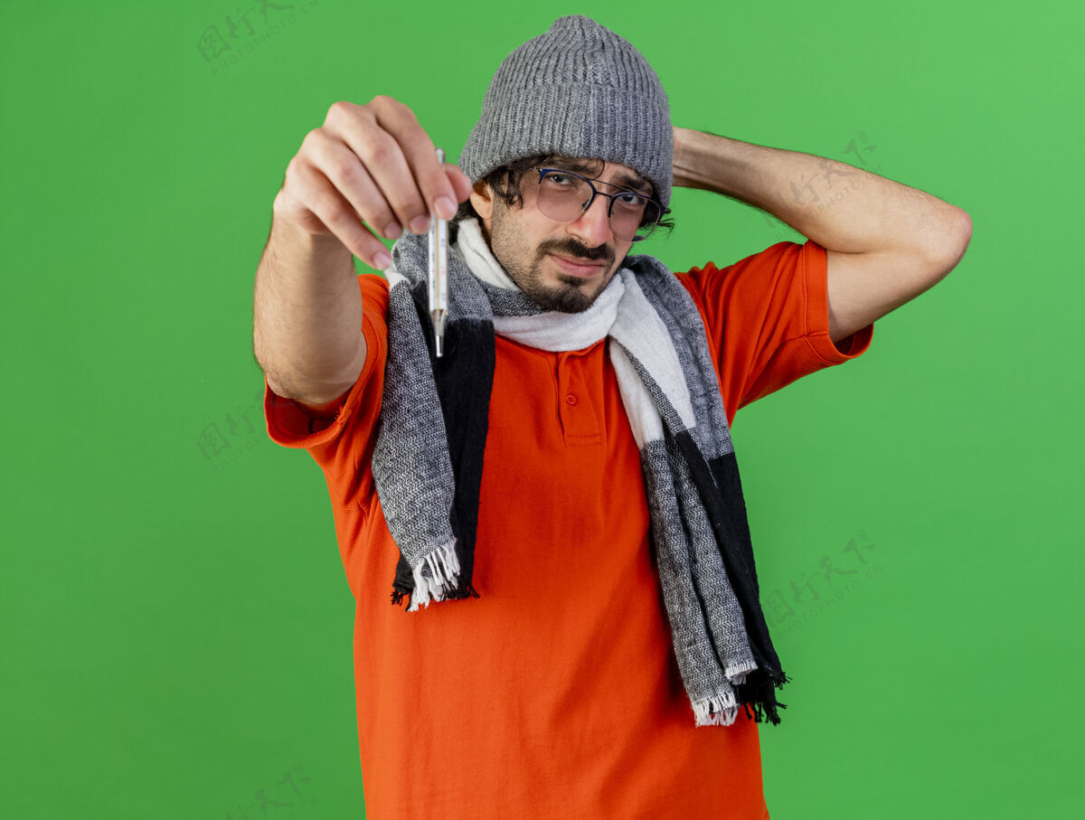 衣服皱眉的年轻病人戴着眼镜 戴着冬天的帽子 戴着围巾 把体温计伸到前面 手放在脑后 看着前面 隔离在绿色的墙上 留着复印空间人体温计眼镜
