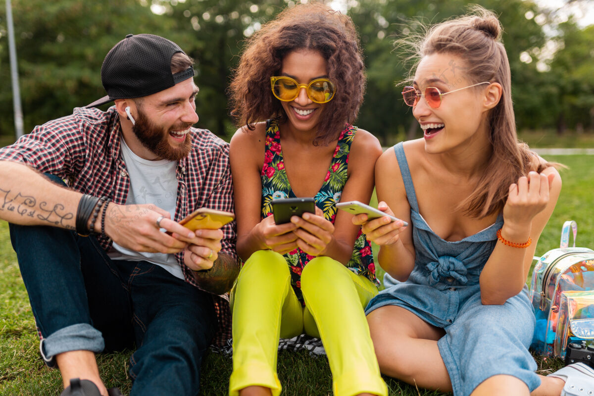 朋友快乐的年轻朋友们坐在公园里用智能手机微笑 男人和女人一起玩手机太阳镜积极