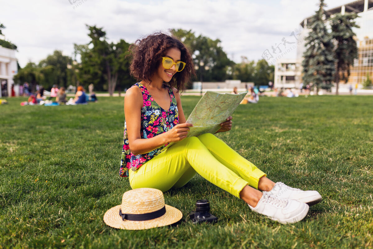 魅力年轻时尚的黑人女子在公园享受夏日时尚的乐趣 五颜六色的时髦装扮 坐在草地上 旅行者拿着地图和草帽明亮年轻卷曲