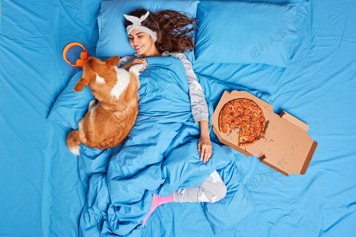 家庭高兴的年轻黑发女子玩狗穿着舒适的睡衣懒得起床吃美味的比萨饼忘记所有的工作放松和最喜欢的宠物一起睡好觉毯子家庭休息