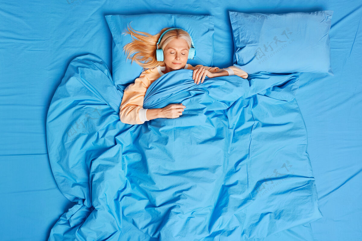 休息放松的金发中年妇女睡在舒适的床上 用无线耳机听音乐宁静放松房子