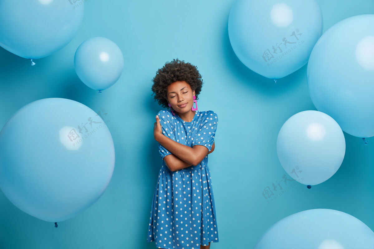 女人高兴的黑皮肤女人拥抱自己 高兴地闭上眼睛 摆出蓝色气球的姿势庆祝服饰室内