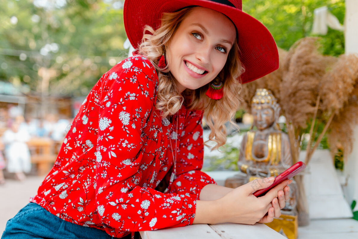 外表迷人的时尚金发微笑的女人在稻草红帽子和衬衫夏季时尚服装举行使用智能手机咖啡厅情感电话年轻