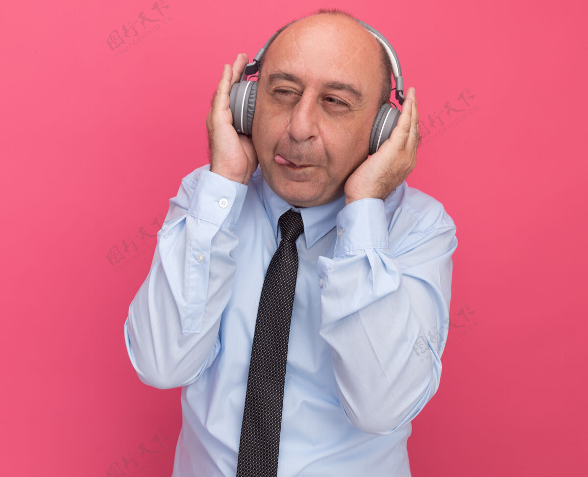 耳机贪婪地看着身边的中年男子 穿着白色t恤 打着领带 把耳机孤立在粉红色的墙上中年男人穿