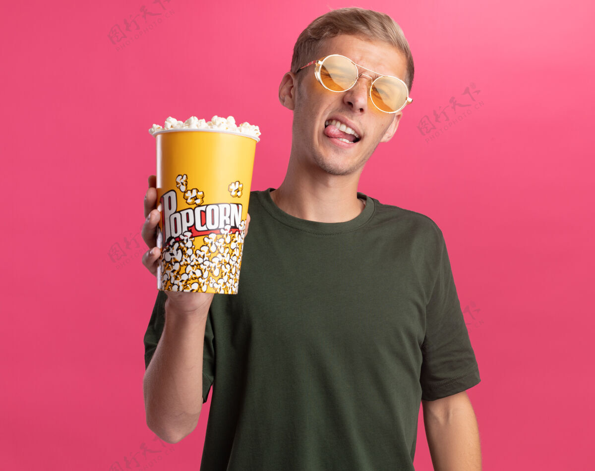 水桶快乐的年轻帅哥穿着绿色衬衫 戴着眼镜 手里拿着一桶爆米花 在粉红色的墙上露出孤立的舌头舌头眼镜衬衫
