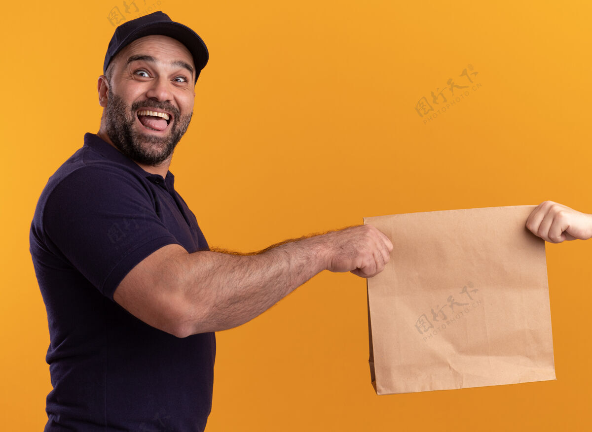 帽子兴奋的中年送货员穿着制服 戴着帽子 把纸食品包装交给隔离在黄色墙上的客户纸张包装中年