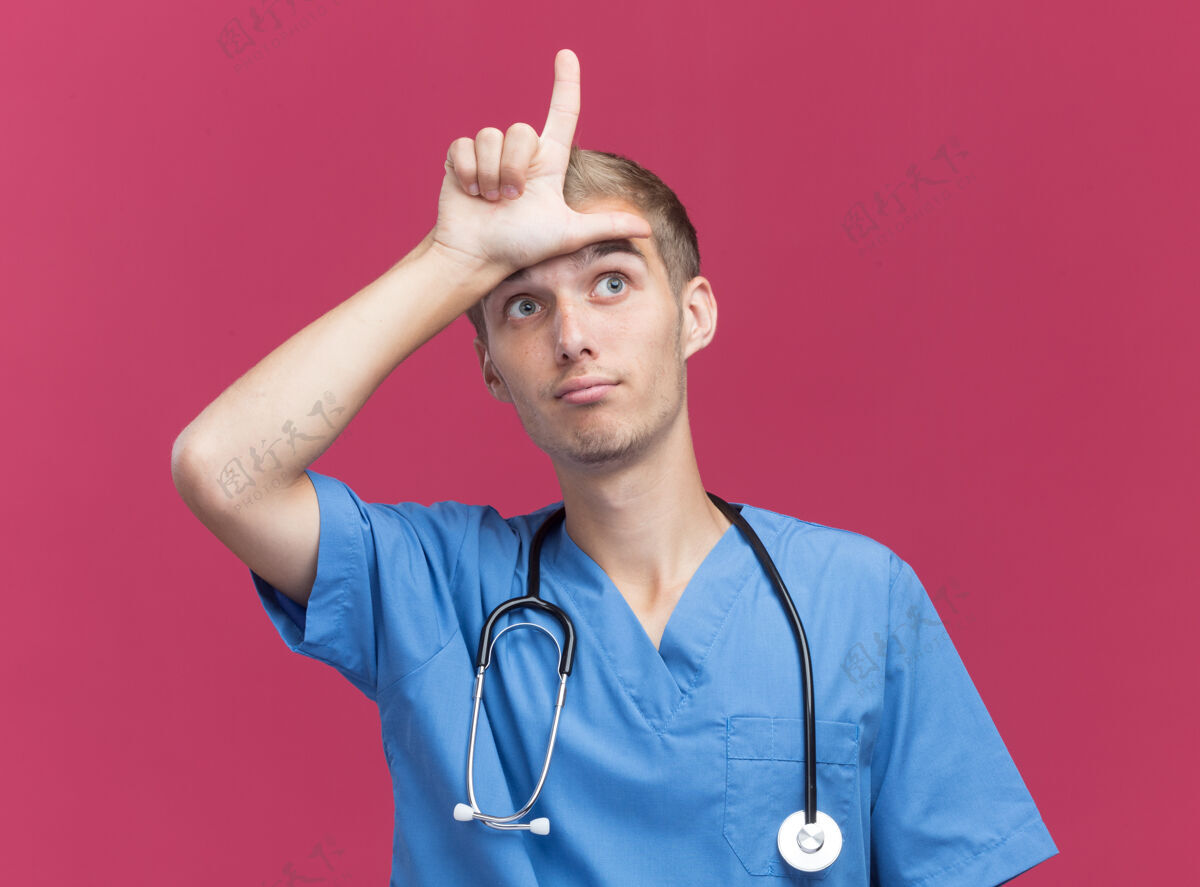 听诊器令人印象深刻的年轻男医生穿着医生制服 听诊器显示失败的姿态隔离在粉红色的墙上手势穿着表演