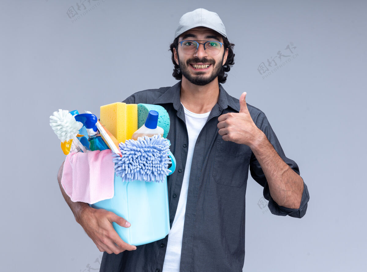 T恤微笑着的年轻帅气的清洁工 穿着t恤 戴着帽子 手里拿着一桶清洁工具 在白色的墙上孤立地竖起大拇指显示清洁水桶