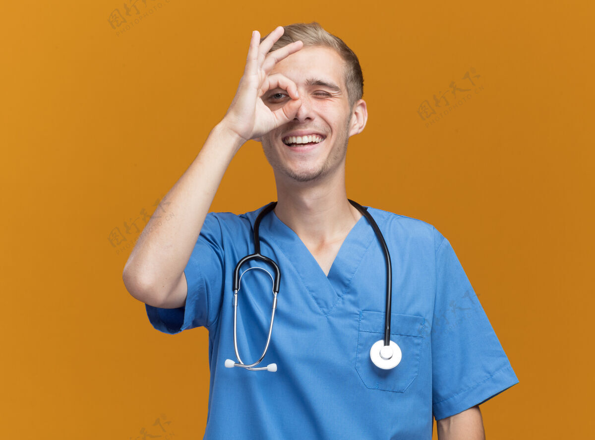 年轻微笑着的年轻男医生穿着医生制服 带着听诊器 在橙色的墙上显示出孤立的表情和姿势展示微笑医生