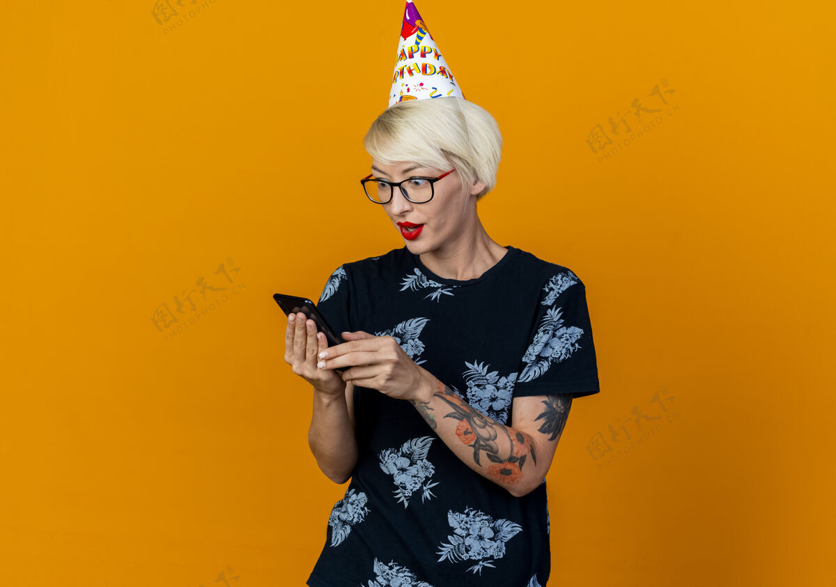 电话令人印象深刻的年轻金发派对女孩戴着眼镜和生日帽举行 看着手机孤立的橙色背景与复制空间帽子复制年轻