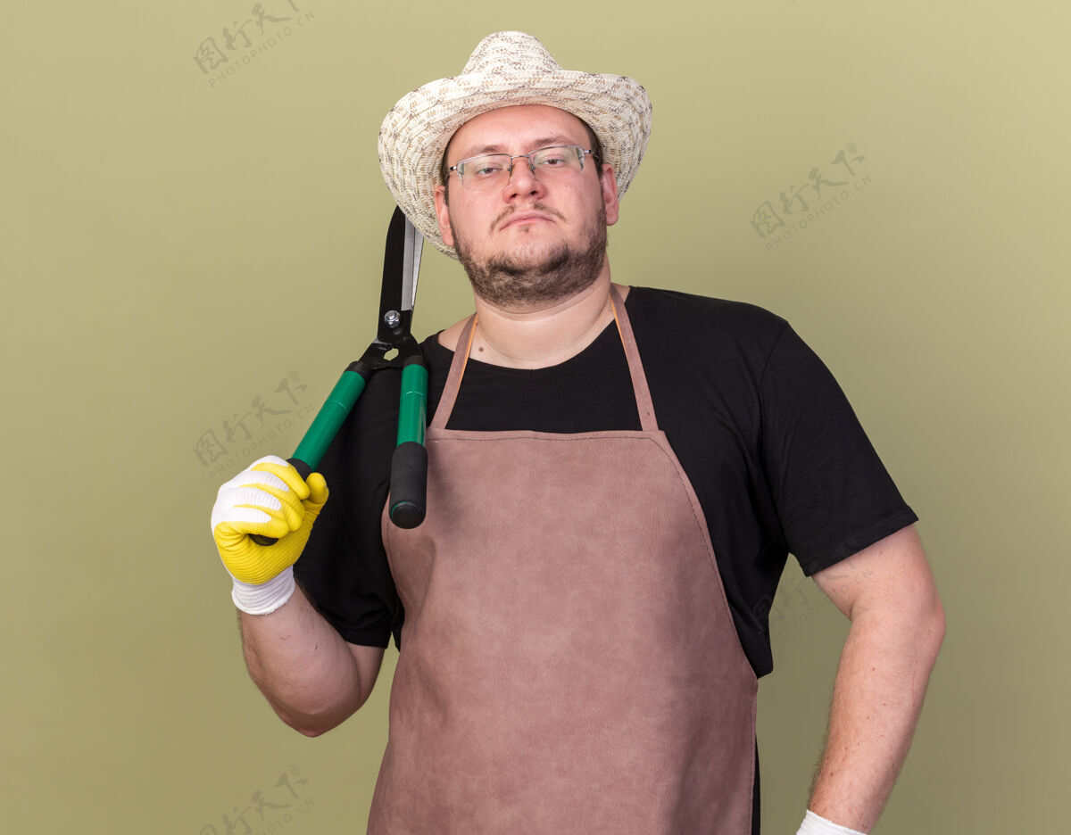 持有自信的年轻男性园丁戴着园艺帽和手套 肩上拿着剪刀 手放在臀部 隔离在橄榄绿的墙上手套肩膀帽子