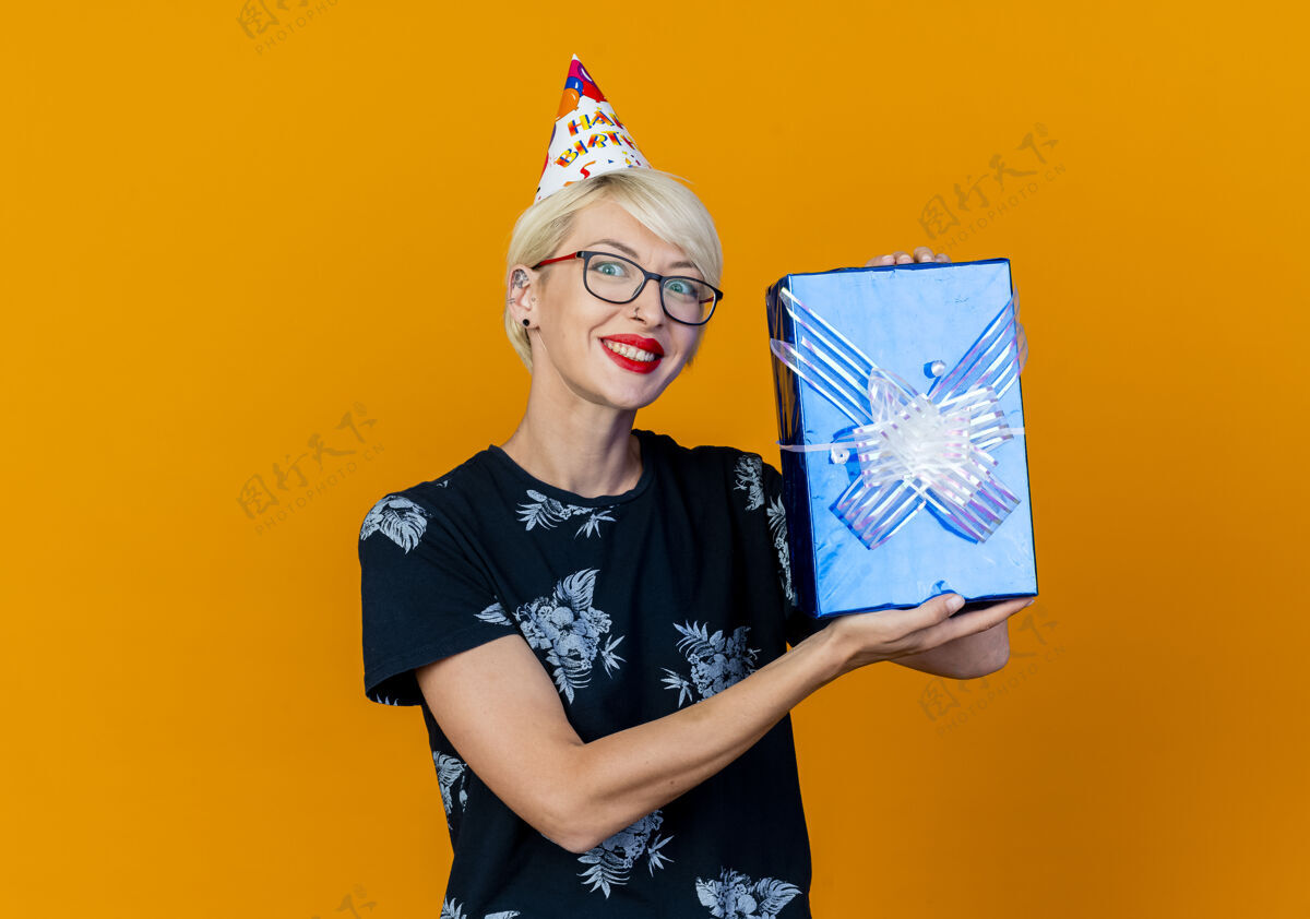 眼镜微笑的金发派对女孩戴着眼镜 戴着生日帽 看着相机 展示着一个孤立的礼物盒 背景是橙色 有复印空间秀盒子年轻