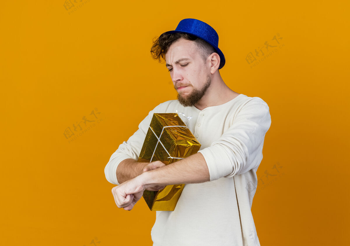 复制皱眉年轻英俊的斯拉夫党人戴着党帽手持礼品盒触摸手腕假装看着手表孤立的橙色背景与复制空间手腕盒子年轻人