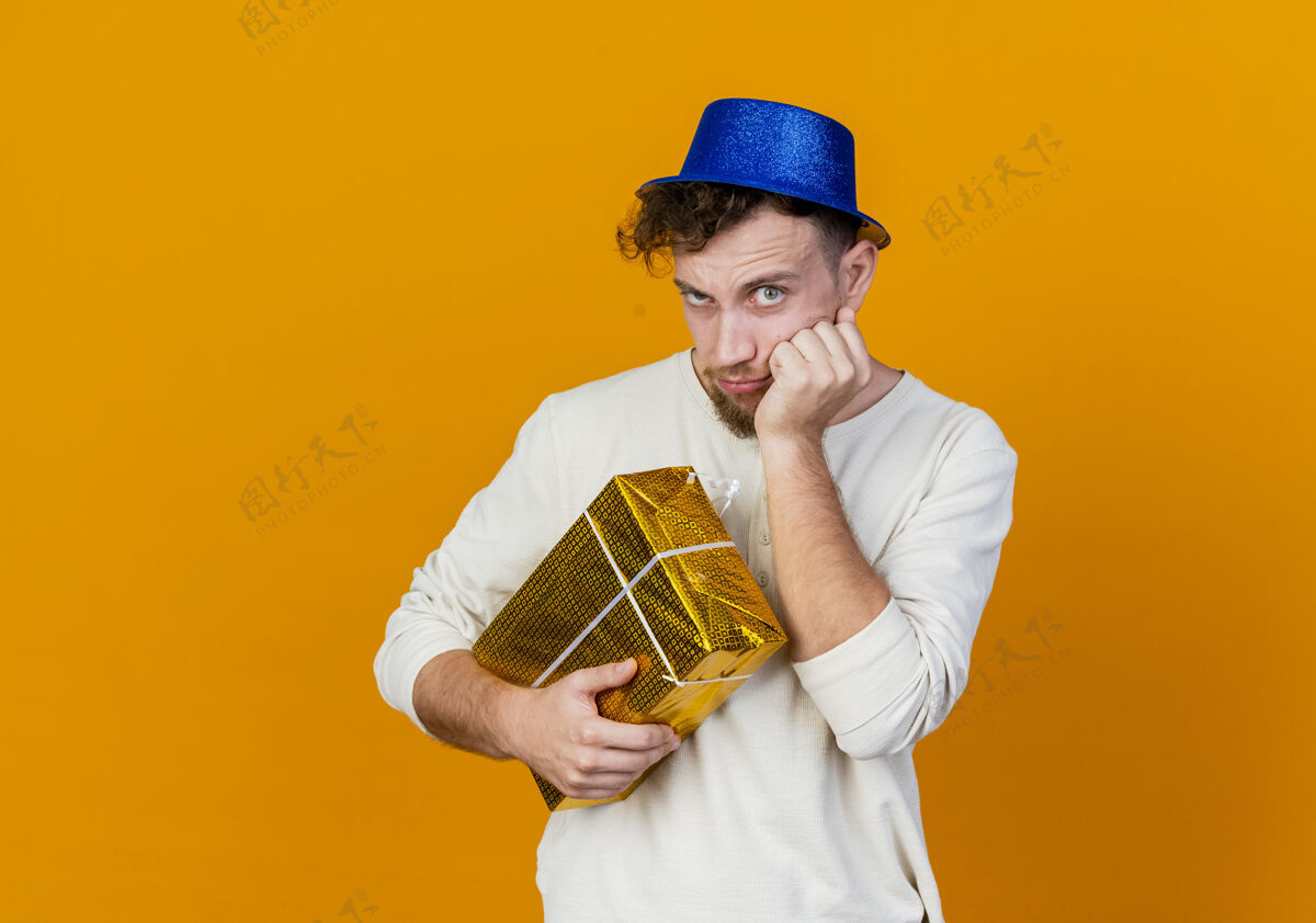 橙色可疑的年轻英俊的斯拉夫党人戴着党的帽子拿着礼品盒把手放在脸上看着相机孤立在橙色背景与复制空间脸帽子怀疑