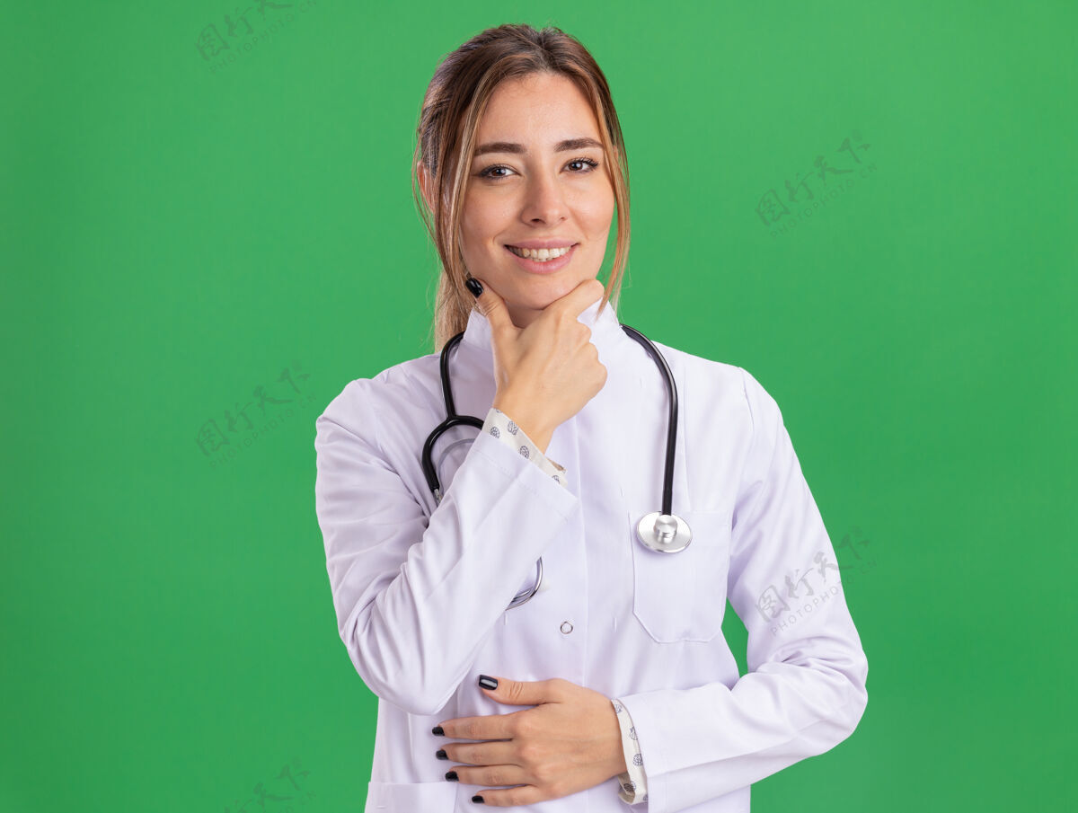 下巴面带微笑的年轻女医生穿着带听诊器的医用长袍 把下巴孤立地抓在绿色的墙上穿医生抓