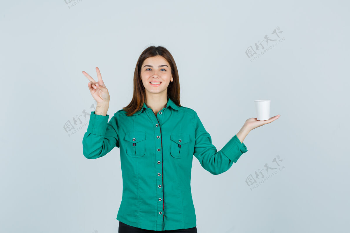 咖啡年轻女士拿着一杯塑料咖啡 穿着衬衫展示胜利标志 看上去很高兴正视图时尚女孩而