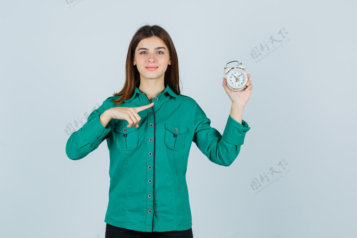 成人穿着衬衫的年轻女士拿着闹钟 指着闹钟 看上去很高兴 前视图眼睛水疗人
