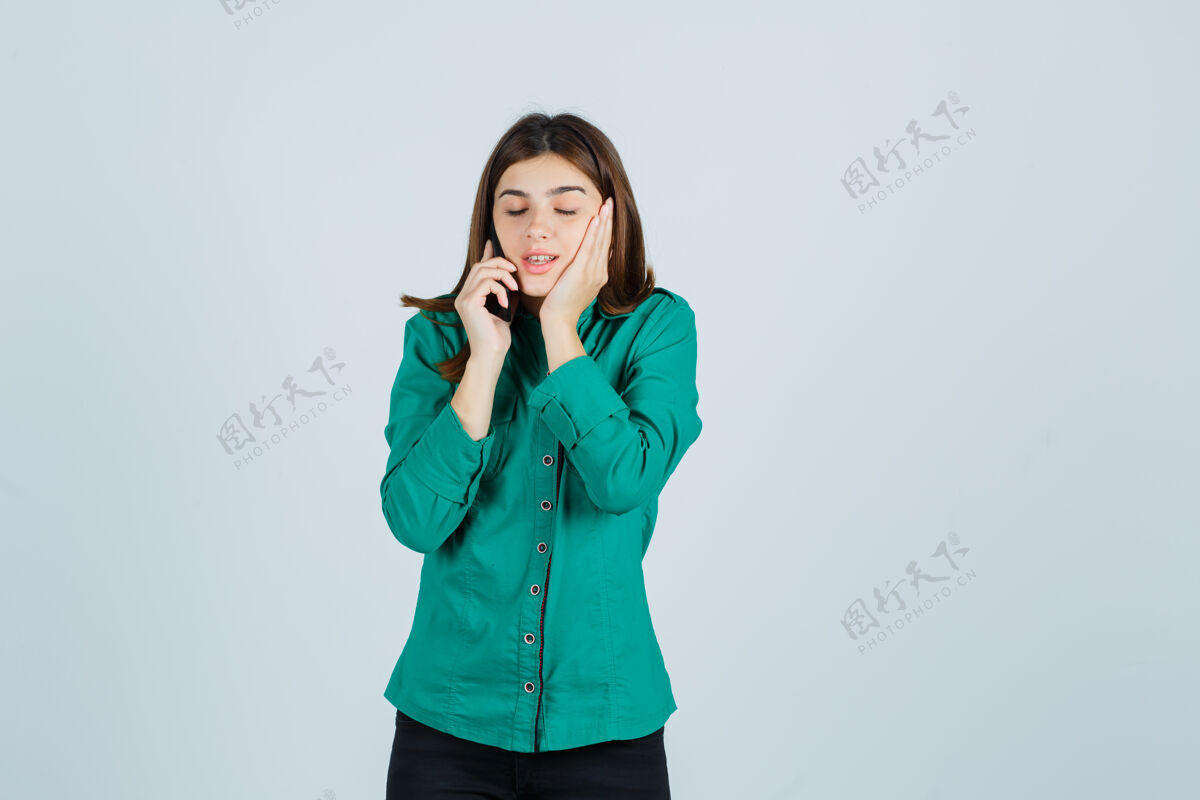 头发年轻的女孩在打电话 手放在脸颊上 穿着绿色上衣 黑色裤子 看起来很好奇 前视图衬衫电话兴趣