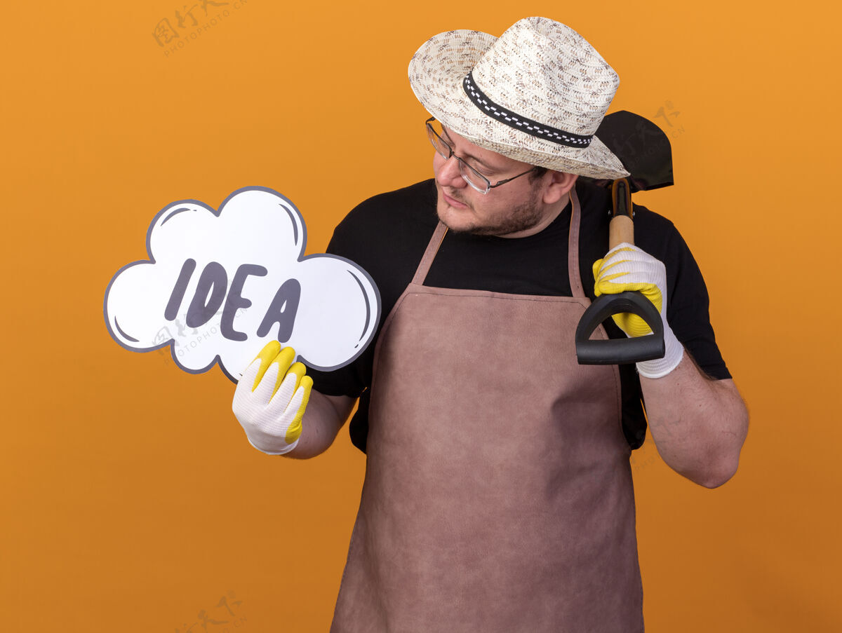 思考年轻的男园丁戴着园艺帽 戴着园艺手套 手里拿着铁锹 看着他手上孤立在橙色墙上的创意泡泡穿泡泡手套