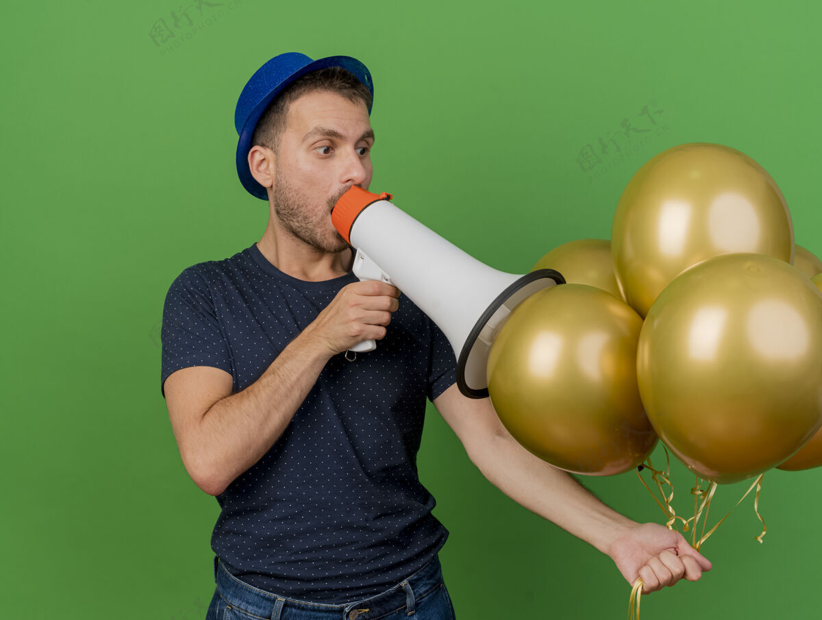 聚会一个戴着蓝色派对帽的英俊白种人拿着氦气球 对着绿色背景上的大喇叭讲话 并留有复印空间举行人气球