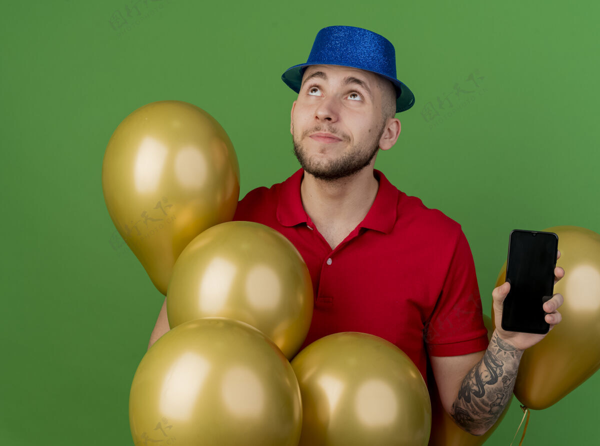 电话体贴的年轻英俊的斯拉夫党人戴着党的帽子站在气球显示手机在绿色背景上看孤立斯拉夫人年轻人气球