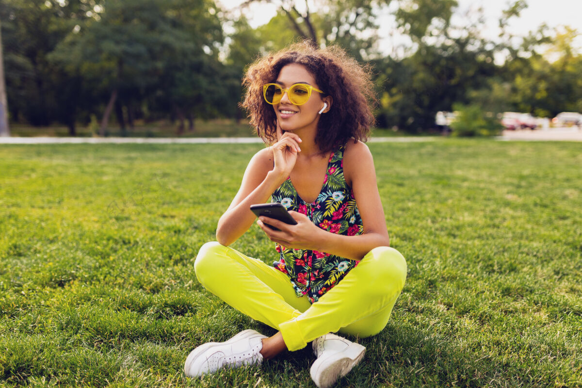 混血儿年轻时尚微笑的黑人女子用智能手机听音乐无线耳机在公园里玩得开心 夏日时尚多彩的风格 坐在草地上 戴着黄色太阳镜耳机享受音乐
