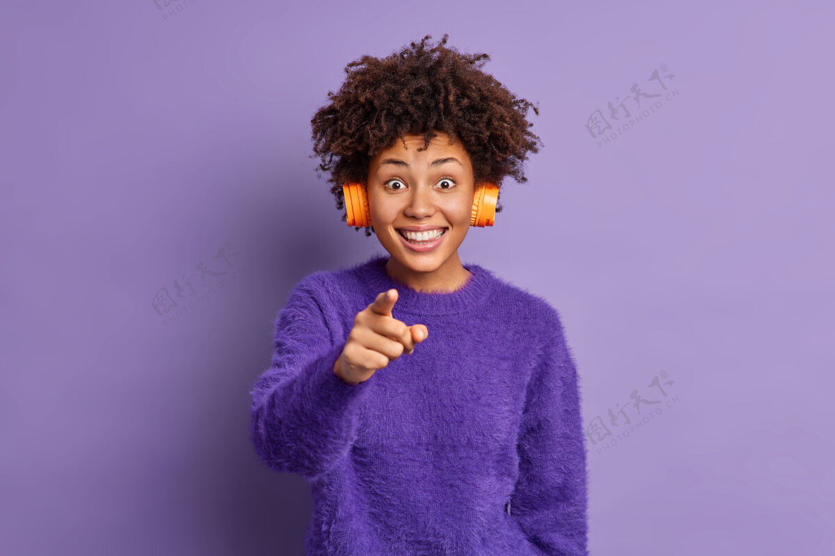 耳机照片中开朗的非洲裔美国女人开心地笑着 直指镜头感到很高兴选择你戴着耳机在耳朵上听喜欢的音乐牙齿乐观头发