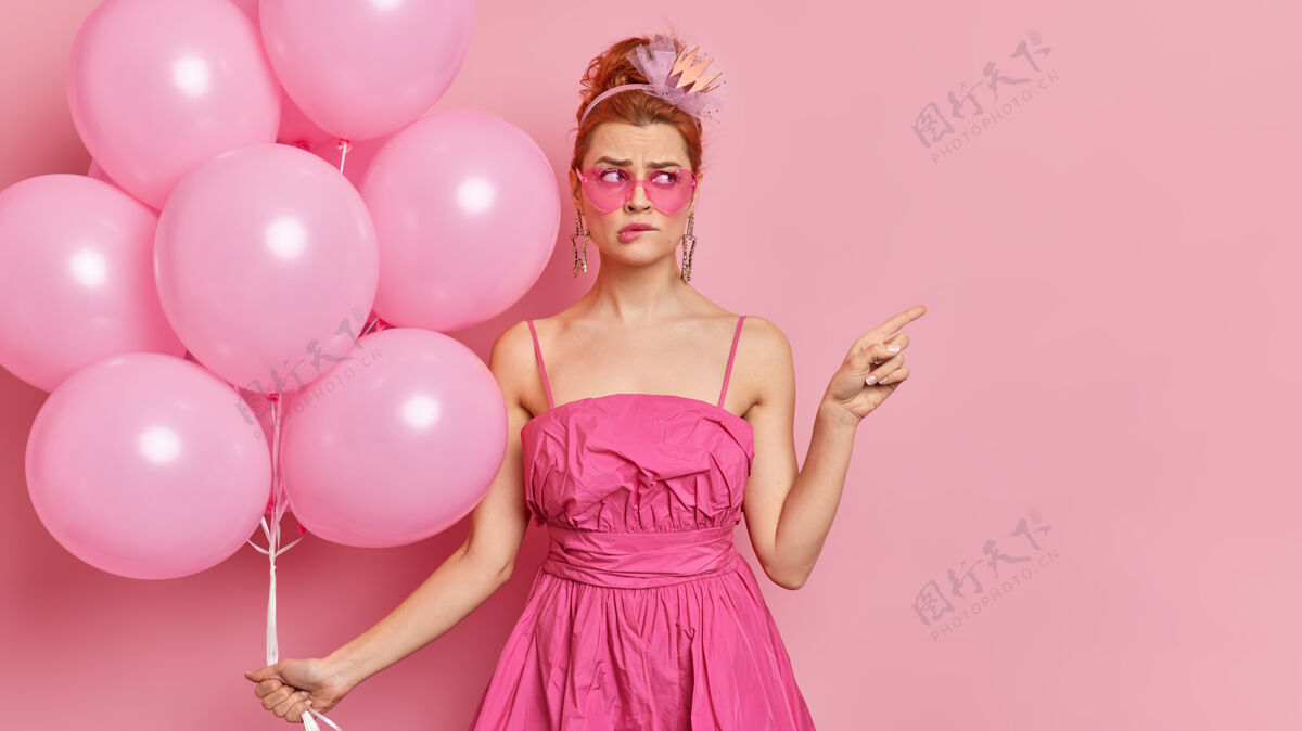 罗西迷惑不解的红发年轻女子穿着迷人的服装 穿着粉色的衣服 在母鸡派对上摆出充气气球的姿势 在空位上点着 咬着嘴唇 展示你广告内容的位置复古充气漂亮