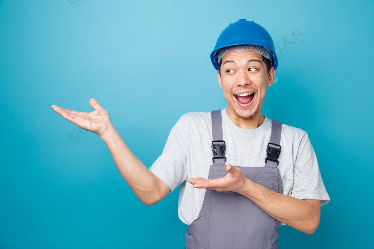 穿兴奋的年轻建筑工人戴着安全帽和制服 用手看着并指着旁边头盔指向工人