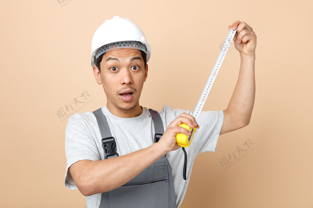 持有年轻的建筑工人戴着安全帽 穿着制服拿着卷尺奶油制服安全