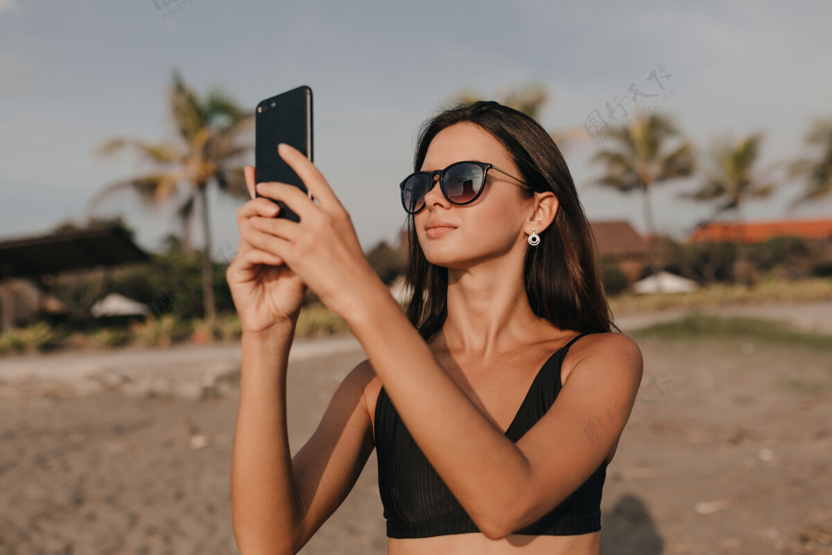 放松时尚可爱的女人戴着墨镜深色头发在沙滩上用智能手机拍照散步女人快乐